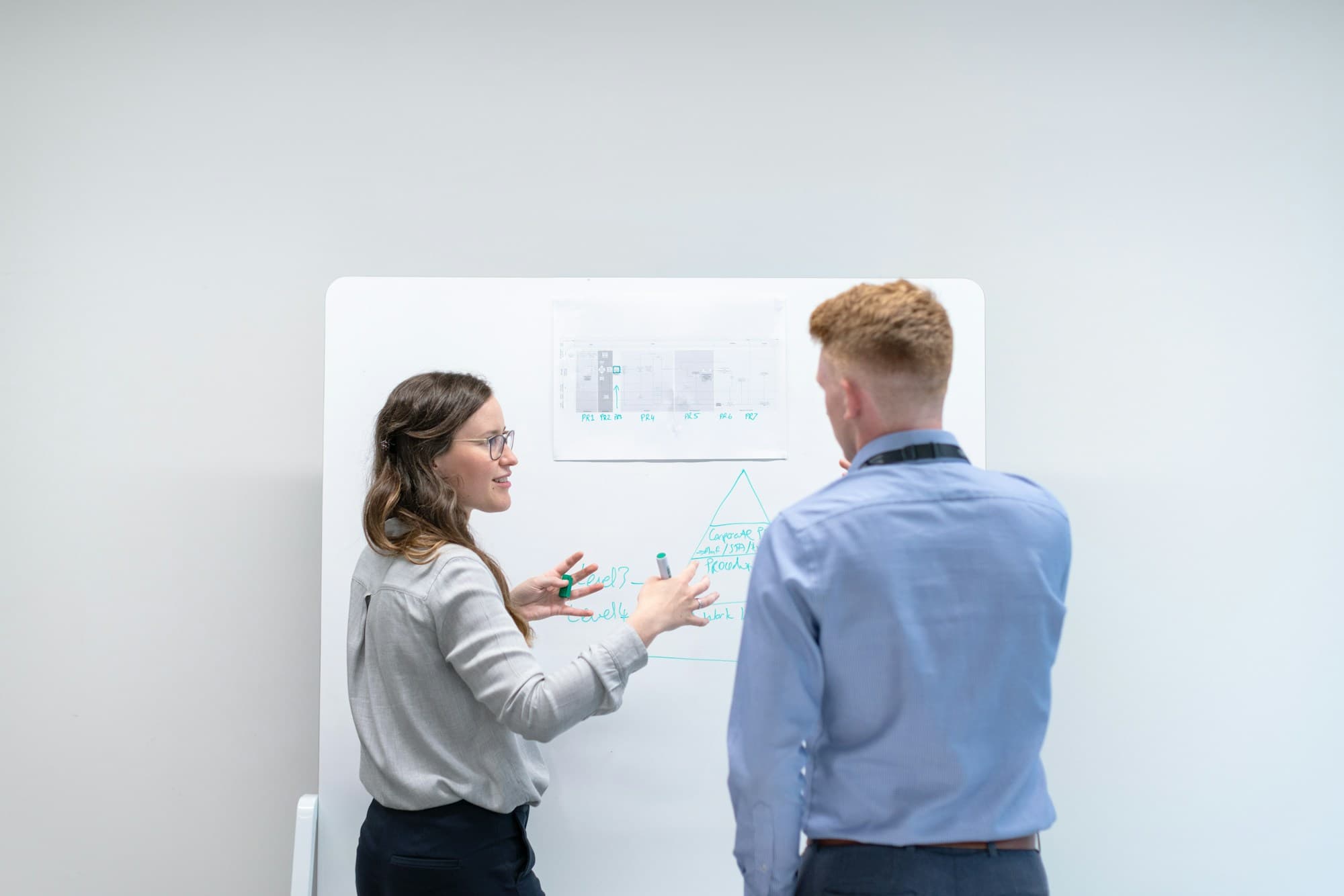 A colleague explains a diagram on a whiteboard, marker in hand, while another listens with their back to camera.