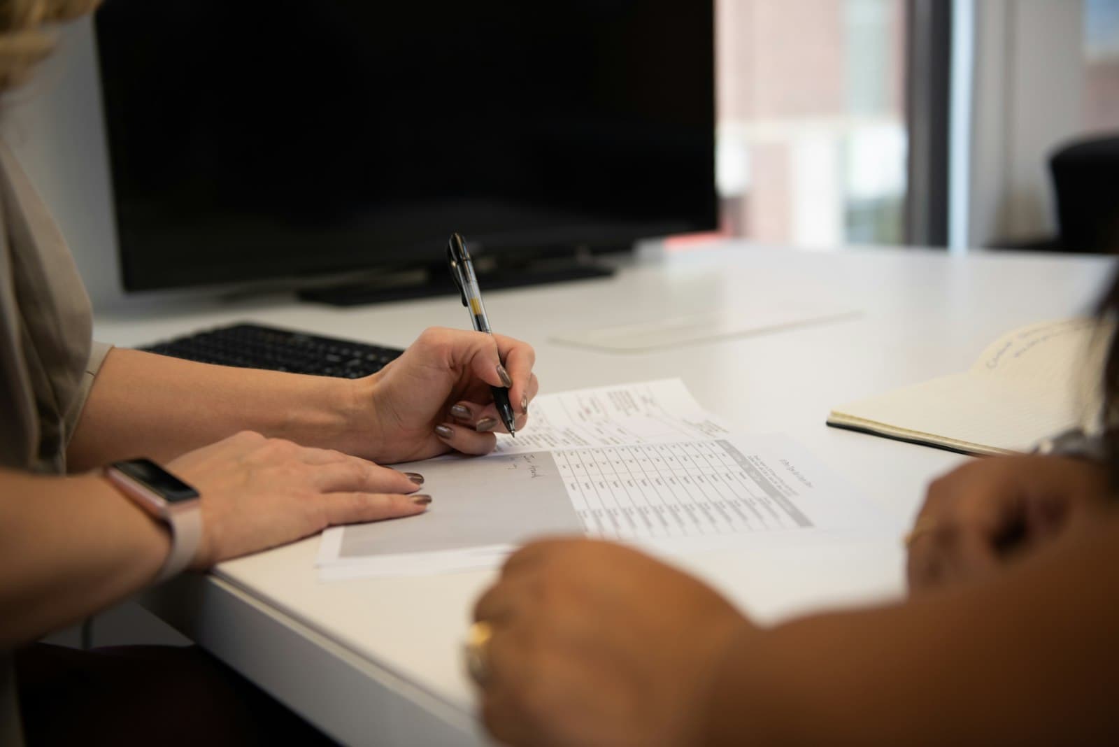 Two people at a desk, one pointing to a printed document while the other writes notes.