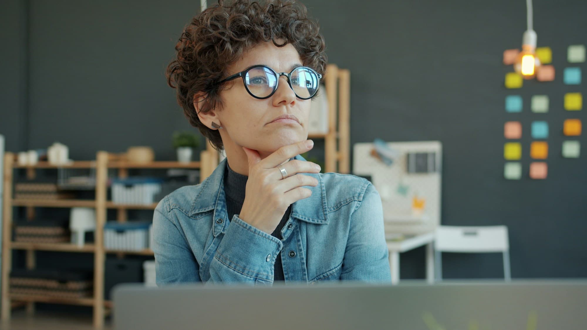 A woman rests her chin on her hand, considering something thoughtfully, laptop open in front of her.
